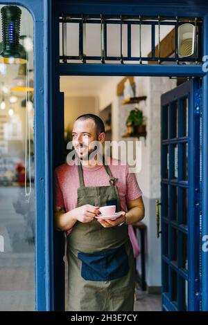 Positiver bärtiger Barista auf der Schürze mit einer Tasse heißen Kaffee in den Händen, der vor dem Eingang des modernen Cafés steht Stockfoto