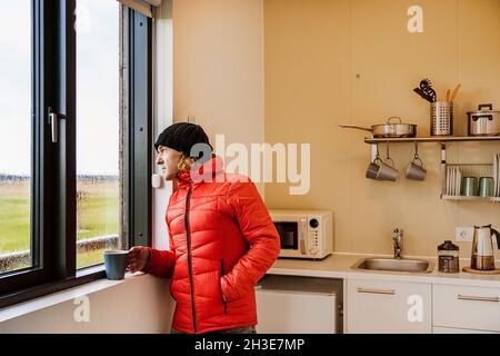 Ruhiger junger männlicher Reisender in warmer roter Jacke und Hut, trinkend Tasse heißen Kaffee und Blick aus dem Fenster stehend in der Küche im Haus an regnerischen Tag in Cou Stockfoto