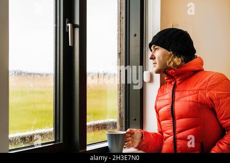 Ruhiger junger männlicher Reisender in warmer roter Jacke und Hut, trinkend Tasse heißen Kaffee und Blick aus dem Fenster stehend in der Küche im Haus an regnerischen Tag in Cou Stockfoto