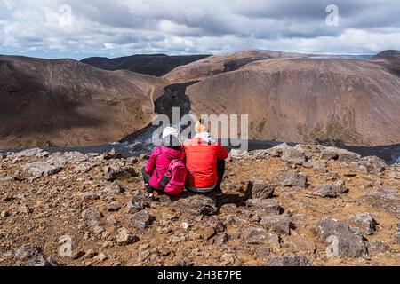 Rückansicht eines unverkennbaren Paares in warmer Oberbekleidung, das auf felsigem Boden sitzt und die malerische Landschaft des aktiven Vulkans Fagradalsfjall in Icel bewundert Stockfoto