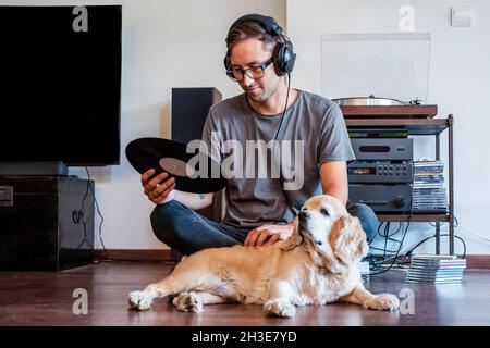 Mann, der Kopfhörer mit dem Player verbindet, während er auf dem Boden mit Cocker Spaniel Welpe in hellem Raum sitzt Stockfoto