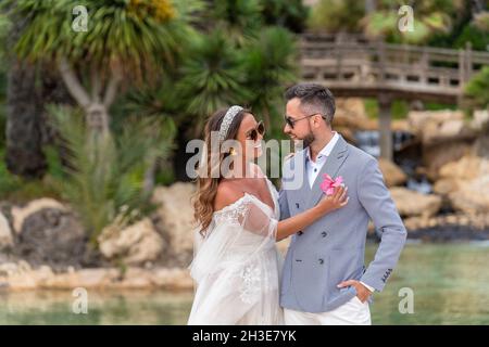 Lächelndes Ehepaar in Hochzeitskleidung und Sonnenbrille, das in der Nähe von See und grünen Bäumen mit Pflanzen steht, während sie einander ansieht und Frau m gibt Stockfoto