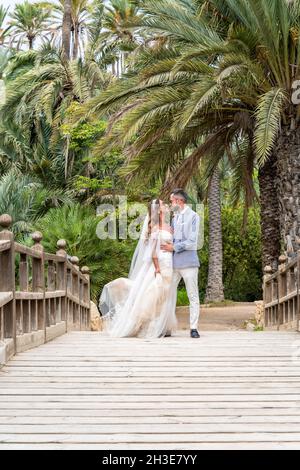 Verheiratetes Paar in Hochzeitsoutfits, das auf einem hölzernen Steg mit Geländer steht, während es umarmt und sich in der Nähe von grünen Palmen und Pflanzen in g anschaut Stockfoto