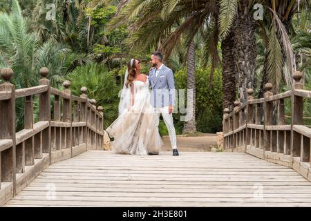 Verheiratetes Paar in Hochzeitsoutfits, das auf einem hölzernen Steg mit Geländer steht, während es umarmt und sich in der Nähe von grünen Palmen und Pflanzen in g anschaut Stockfoto