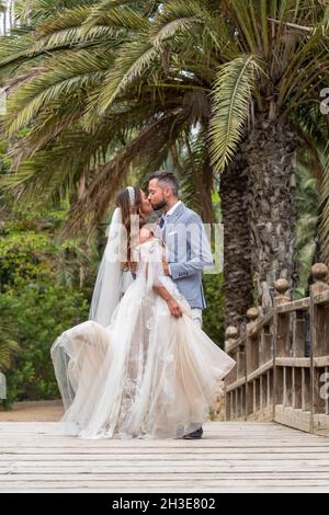 Verheiratetes Paar in Hochzeitskleidung, das am Sommertag auf einem Holzsteg mit Geländer steht und sich in der Nähe von grünen Palmen und Pflanzen im Garten küsst Stockfoto