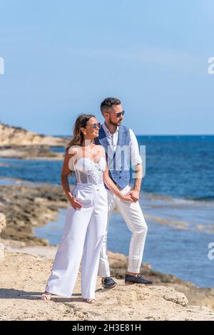 Ganzer Körper eines jungen Paares in elegantem Outfit und Sonnenbrille, die in der Nähe des Meeres auf einem Hügel an der felsigen Küste steht, während sie die Hände hält und unter blauem Himmel wegschaut Stockfoto