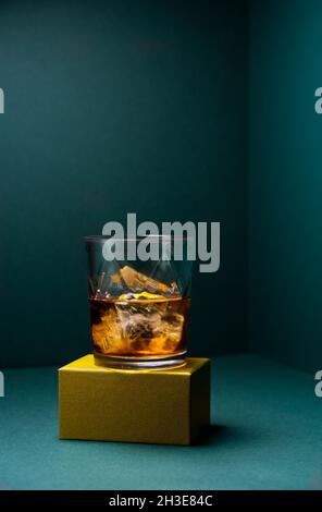 Vorderansicht einer Glasschale mit kaltem Whiskey und Eiswürfeln, die auf einer goldfarbenen, robusten Basis und einem türkisblauen Eckhintergrund platziert ist Stockfoto