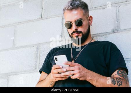 Glücklicher bärtiger Kerl mit Tattoos in schwarzem T-Shirt und Sonnenbrille, der an der Wand des Gebäudes steht und ein Smartphone bei Tageslicht benutzt Stockfoto