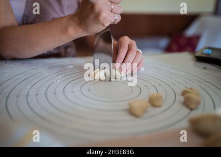 Hoher Winkel der Ernte anonyme Dame schneiden rohen Teig mit Kitt Messer während der Vorbereitung hausgemachte Knödel sitzen am Tisch in der Küche Stockfoto