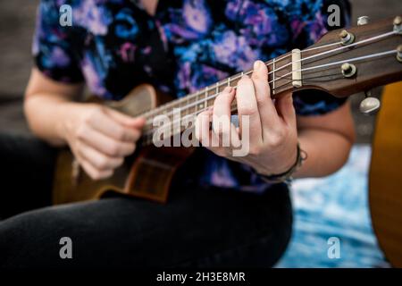 Beschnittene, nicht erkennbare, talentierte Musikerin mit braunen Haaren in legerer Kleidung, die Ukulele spielt und Gesang singt, während sie am Sandstrand in Nat sitzt Stockfoto