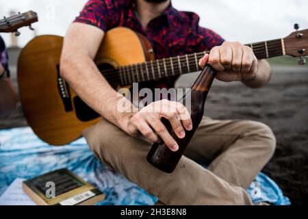 Von unten zeigt ein nicht erkennbarer Musiker, der mit Akustikgitarre sitzt und eine Flasche Bier öffnet, während er Zeit am Strand in der Natur in d verbringt Stockfoto