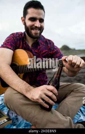 Von unten ein positiv bärtiger Musiker, der mit Akustikgitarre sitzt und eine Flasche Bier öffnet, während er am Tag Zeit am Strand in der Natur verbringt Stockfoto