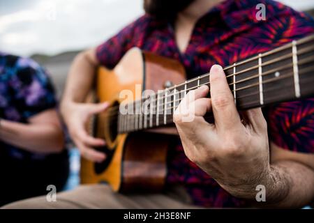 Cropped unkenntlich talentierte machen Musiker in lässigen Kleidern spielen akustische Gitarre und Gesang, während sie am Sandstrand in der Natur am Tag sitzen Stockfoto