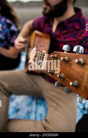 Cropped unkenntlich talentierte machen Musiker in lässigen Kleidern spielen akustische Gitarre und Gesang, während sie am Sandstrand in der Natur am Tag sitzen Stockfoto