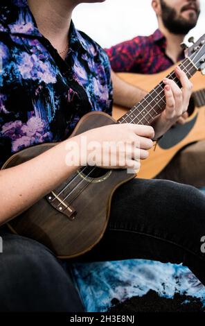 Beschnittene, nicht erkennbare, talentierte Musikerin mit braunen Haaren in legerer Kleidung, die Ukulele spielt und Gesang singt, während sie am Sandstrand in Nat sitzt Stockfoto