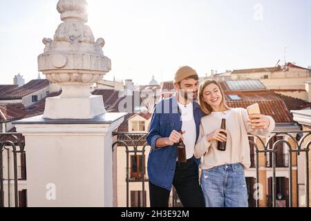 Positive junge Freunde, die zusammen Selfie machen, während sie Bier aus Glasflaschen trinken und sich am Zaun auf dem Balkon lehnen Stockfoto