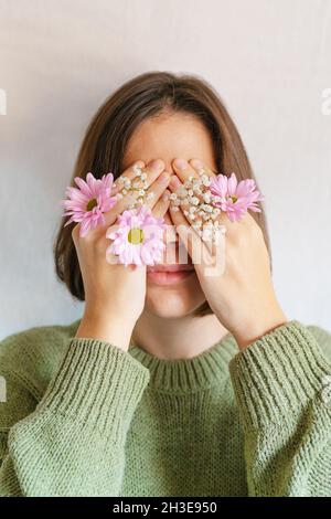 Junge Frau in Strickpullover versteckt Augen mit Händen und blühenden Chrysantheme und Gypsophila Blumen vor grauem Hintergrund Stockfoto