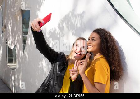 Seitenansicht glücklicher junger multiethnischer Freundinnen, die lächeln und zwei Finger zeigen, während sie Selfie auf der Straße machen Stockfoto