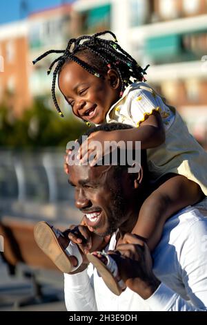 Glückliches afroamerikanisches Mädchen mit dunklen Zöpfen, das auf den Schultern eines fröhlichen Vaters sitzt und beim Springen auf der Straße im Sonnenlicht Spaß hat Stockfoto