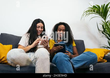 Junge, vielfältige Freundinnen in legerer Kleidung lächeln, während sie auf dem Sofa sitzen und im Wohnzimmer zu Hause auf dem Smartphone surfen Stockfoto