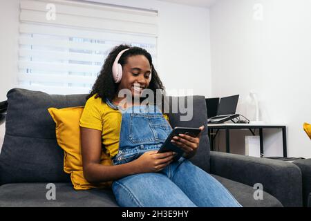 Von unten eine glückliche afroamerikanische Frau in stilvollem Outfit, die in Kopfhörern Musik hört und Tablet benutzt, während sie auf einem grauen Sofa in hellem Ro sitzt Stockfoto