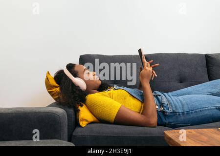 Junge afroamerikanische Frau mit lockigem Haar in Kopfhörern und legerer Kleidung, die auf einem bequemen grauen Sofa liegt und im hellen Raum auf dem Smartphone stöbert Stockfoto