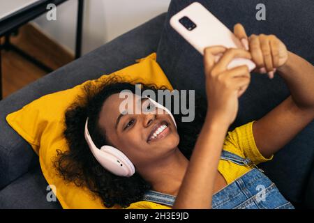Von oben eine junge afroamerikanische Frau mit lockigem Haar in Kopfhörern und legerer Kleidung, die auf einem bequemen grauen Sofa liegt und Selfie auf Smart nimmt Stockfoto