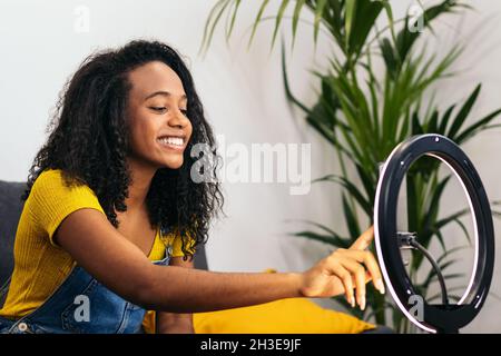 Afroamerikanische Frau in Denim-Kleidung lächelt, während sie auf dem Sofa sitzt und das Smartphone auf einer glühenden Ringlampe berührt Stockfoto