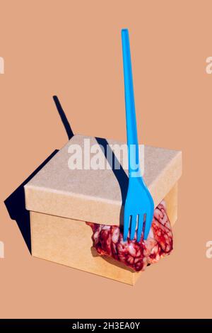 Rohe Gehirne kommen aus Karton mit Deckel und blauer Kunststoffgabel auf orangefarbenem Hintergrund im Studio mit Sonnenlicht Stockfoto