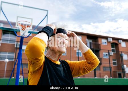 Selbstbewusste, reife Frau im aktiven Ohr und Stirnband, die beim Training auf der Straße mit dem Gebäude in der Nähe des Basketballkorps auf dem Sportplatz steht Stockfoto