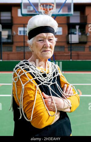 Seitenansicht Naturweibchen im aktiven Ohr und weißem Netz, die während des Trainings auf dem Sportplatz mit Basketballkorb auf die Kamera schauen Stockfoto