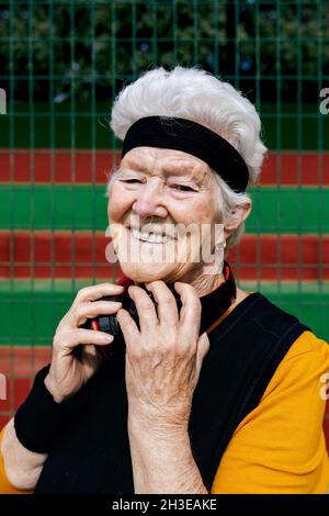 Eine positive, reife Frau mit durchbohrter Nase und Kopfhörern, die Sportkleidung trägt und dabei die Kamera anschaut, während sie während des Trainings auf dem Sportplatz steht Stockfoto