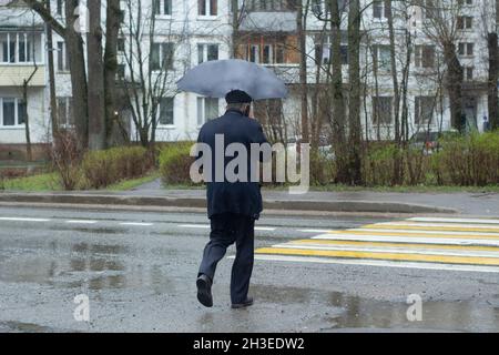 Ein älterer Mann in einem blauen Anzug mit einem Regenschirm überquert die Straße an einer Fußgängerüberführung, ein Blick von hinten. Ein regnerischer Tag. Verkehr. Lifestyle in der Stadt. Stockfoto