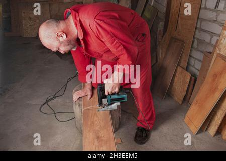 Ein Tischler in einer Werkstatt sägt ein Laminat mit einer Stichsäge. Arbeiter in roten Overalls mit einem Elektrowerkzeug schneidet Parkett. Der Arbeiter schneidet die Bretter mit einem elektrischen j Stockfoto