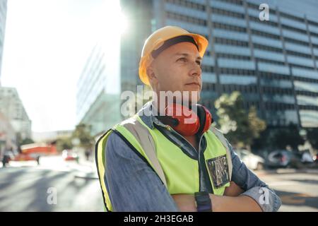 Bauarbeiter schaut nachdenklich weg. Ein mittelerwachsener Arbeiter, der mit gekreuzten Armen in der Stadt steht. Arbeiter mit blauem Kragen, der vor h steht Stockfoto