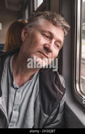 Ein nachdenklicher, gutaussehender Mann mittleren Alters sitzt in der Kutsche eines alten Elektrozuges und schaut aus dem Fenster. Arbeiten Sie in einer anderen Stadt. Ausflüge zwischen Städten Stockfoto
