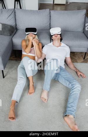 Schockiert Paar in vr Headsets auf dem Teppich neben grauen Couch sitzen Stockfoto