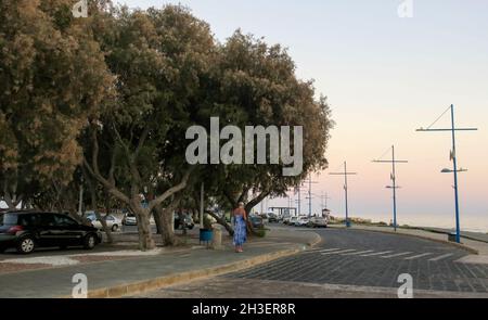Ayia Napa, Zypern - 14. Oktober 2021: Die Straße zum Hafen bei Sonnenuntergang. Auf der linken Seite - ein Parkplatz für Autos, auf der rechten Seite - die Böschung und das Meer, Stockfoto