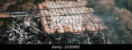 Grillen unter Campingbedingungen. Im Sommer oder Herbst werden Schweinefleischstücke auf dem Grill auf schwelenden Kohlen oder vom Lagerfeuer auf dem Boden gebraten Stockfoto
