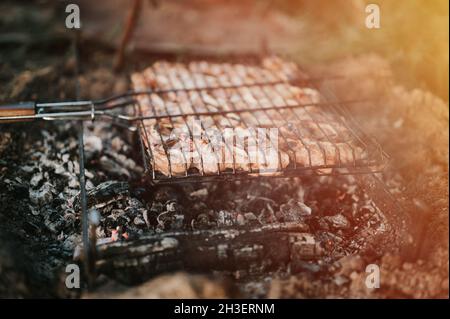 Grill in Campingbedingungen. Stücke von Schweinefleisch mit Zwiebeln werden auf dem Grill auf schwelenden Kohlen oder vom Lagerfeuer auf dem Boden im Sommer oder a gebraten Stockfoto