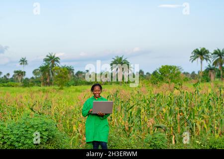 Junge, schön lächelnde afrikanische Bäuerin in der Maisfarm mit einem Laptop an einem sonnigen Tag Stockfoto