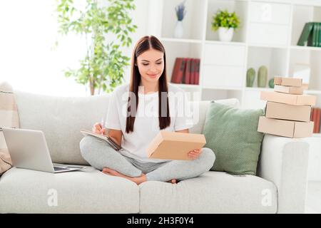 Foto von selbstbewussten positiven Dame halten Papier Paket Pad schreiben Notizen tragen weißes T-Shirt sitzen Couch im Zimmer drinnen Stockfoto