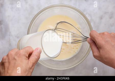 Draufsicht auf eine Frau, die ein Glas Milch in einer Glasschüssel auf einer Marmoroberfläche in gemischte rohe Eier legt Stockfoto