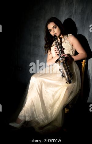 Junge Frau spielt Saxophon. Mädchen in einem weißen flauschigen Kleid posiert mit einem Musikinstrument Stockfoto