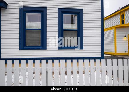 Zwei große blaue Zierfenster an der Außenseite eines weißen Kabeljau-Nebengebäudes mit einem weißen Holzpfostenzaun im Vordergrund. Stockfoto