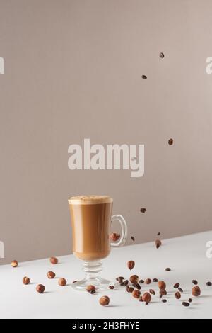 Ein Glas Cappuccino mit duftendem Schaum und fallenden Haselnüssen und gerösteten Kaffeebohnen. Heißer Latte oder Cappuccino in einer Glastasse. Kreatives Design für c Stockfoto