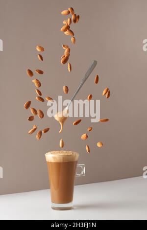 Glasbecher mit Cappuccino und duftendem Schaum und einem Löffel und fallenden Mandeln. Tasse mit Cappuccino, Latte oder Kaffee mit Milch. Kreatives Design für Cafés Stockfoto