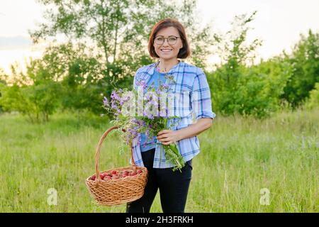 Porträt einer Frau mittleren Alters mit einem Blumenstrauß und einem Korb mit Erdbeeren in der Natur Stockfoto