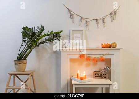 Rahmen mit Schild 'Happy Halloween'. Zamioculcas Pflanzen im Tontopf auf dem Stuhl. Girlande von Skeletten hängt an der Wand. Girlande aus Kunststoff-Kürbissen hängt von Stockfoto