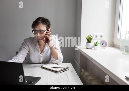 Frau mittleren Alters mit kurzhaariger Brünette in einer Brille arbeitet an einem Laptop in der Nähe des Fensters. Büroangestellte sprechen am Telefon Stockfoto